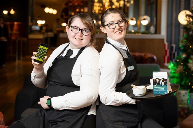 Emma and Kate at work, Emma holding up her phone with AssistiV